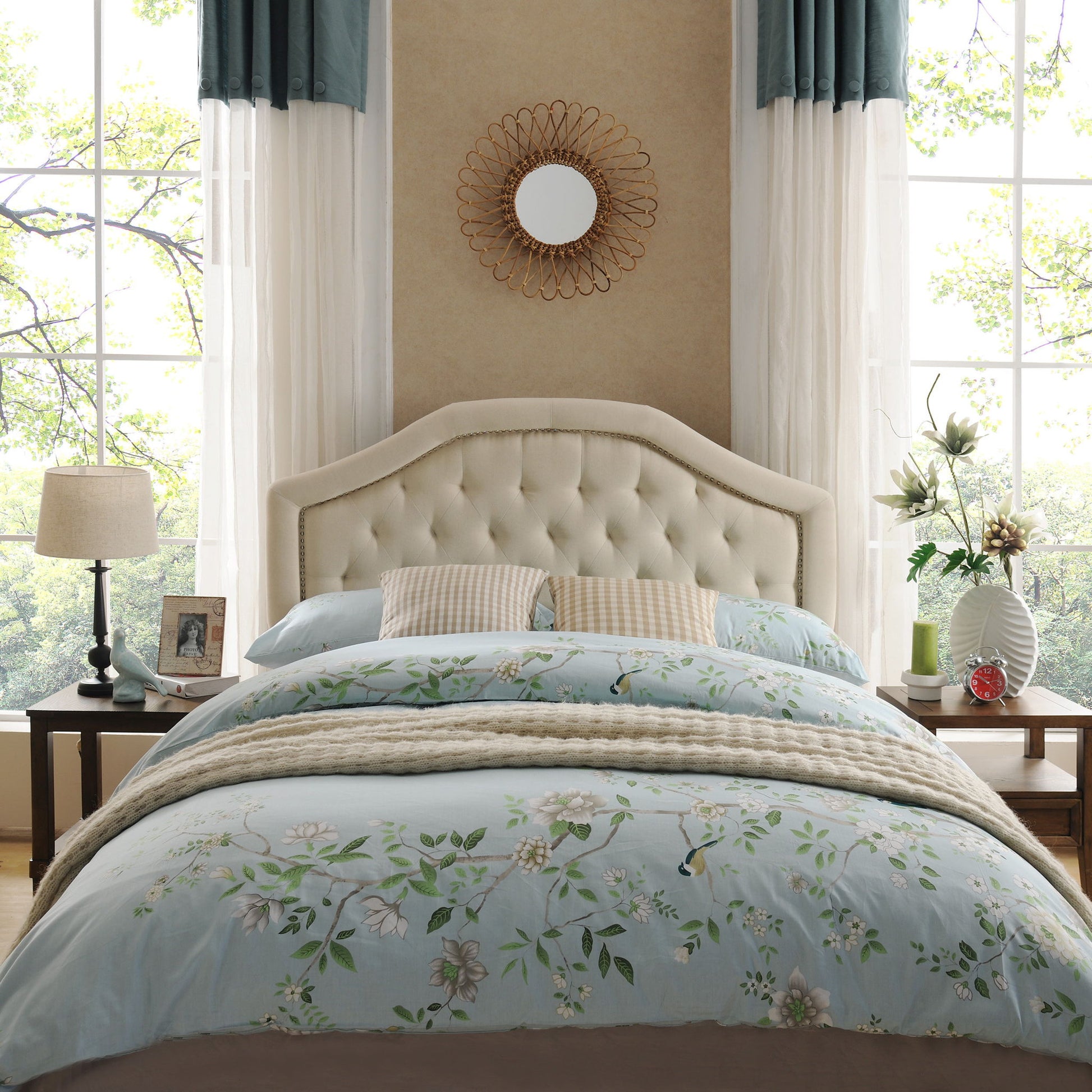 A cozy bedroom featuring the Queen & Full Size Headboard With Diamond Tufting And Nailhead Trim in beige, floral bedding, bedside tables with lamps, framed photos, vases, large windows with green trees outside, and a round mirror above the bed.