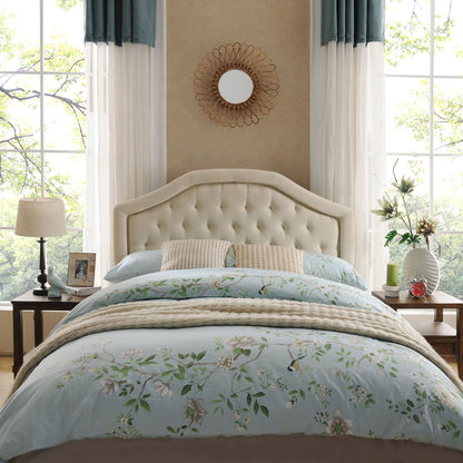 A cozy bedroom featuring the Queen & Full Size Headboard With Diamond Tufting And Nailhead Trim in beige, floral bedding, bedside tables with lamps, framed photos, vases, large windows with green trees outside, and a round mirror above the bed.
