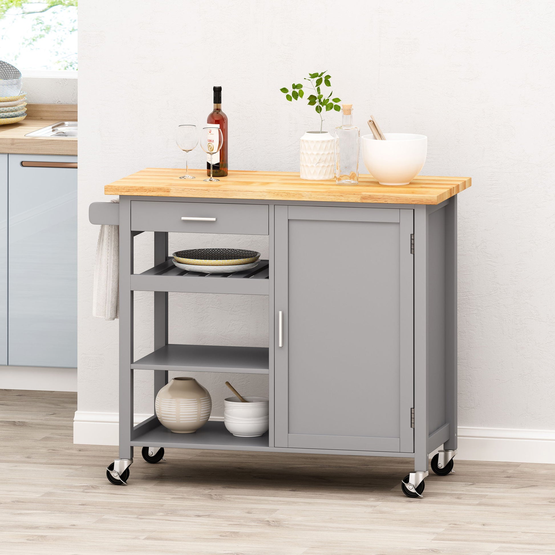 The Contemporary Design Kitchen Cart With Wheels features a gray finish, wooden top, drawer, shelves, and cabinet. Shown adorned with glassware, dishes, a bottle, potted plant, and bowl against a light-colored wall.
