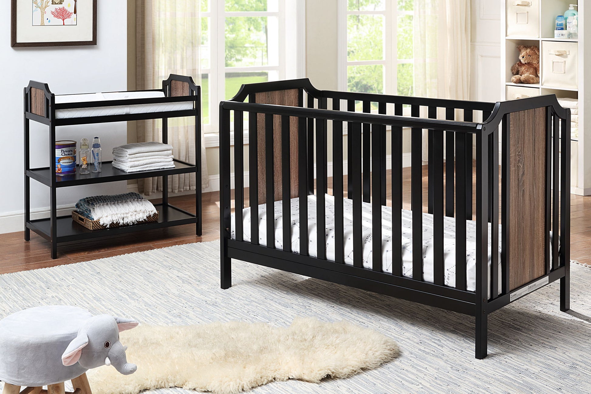 A modern nursery features a black and wood crib, the Brees Changing Table with baby supplies, a white rug, plush elephant stool, shelves of books and toys, and a large window filling the room with natural light.