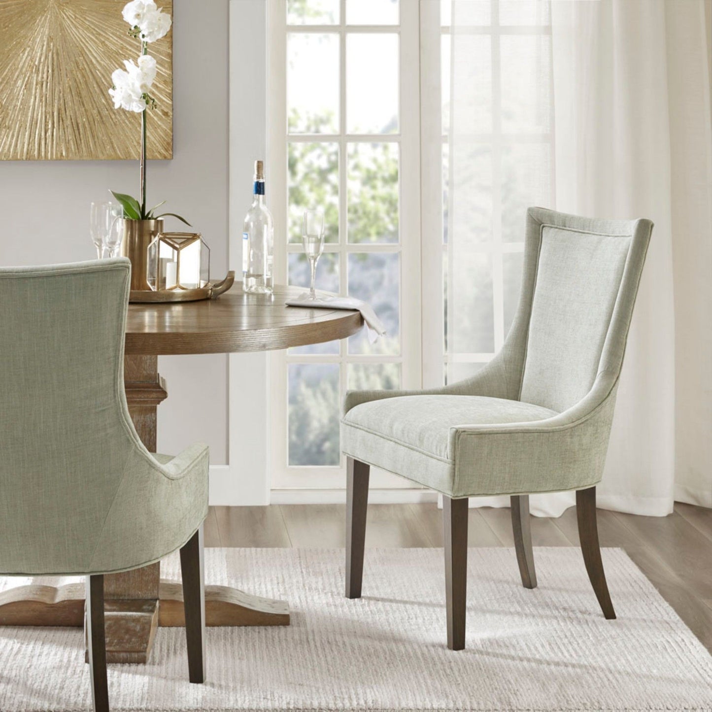 A modern dining room features a round wooden table, an Ultra - Elegant Dining Side Chair set (2, light gray upholstery), a white orchid, and wine by large windows with sheer curtains. Sunlight brightens the neutral decor.