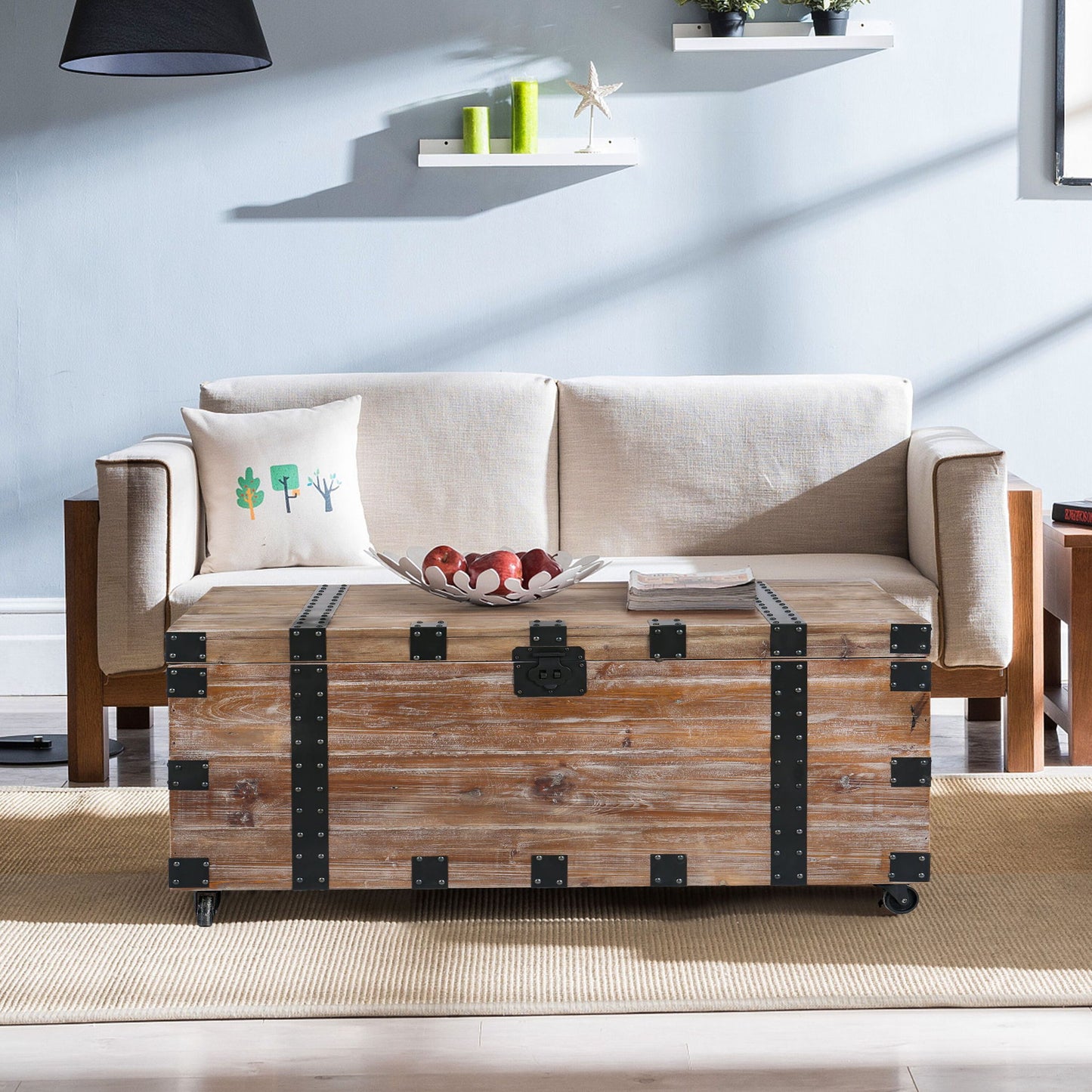 An antique gray trunk table and side table set with large storage dress up the living room. They sit before a light beige sofa with a white pillow, against light blue walls, a beige rug, and floating shelves displaying small decor items.