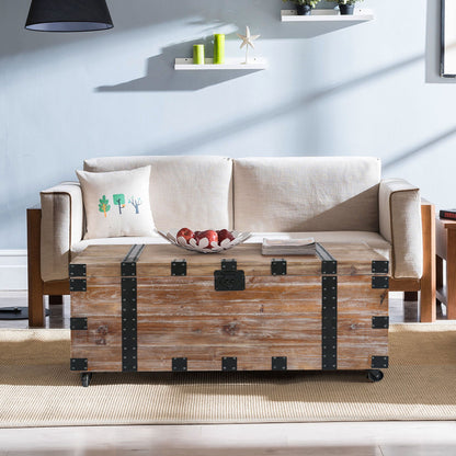 An antique gray trunk table and side table set with large storage dress up the living room. They sit before a light beige sofa with a white pillow, against light blue walls, a beige rug, and floating shelves displaying small decor items.
