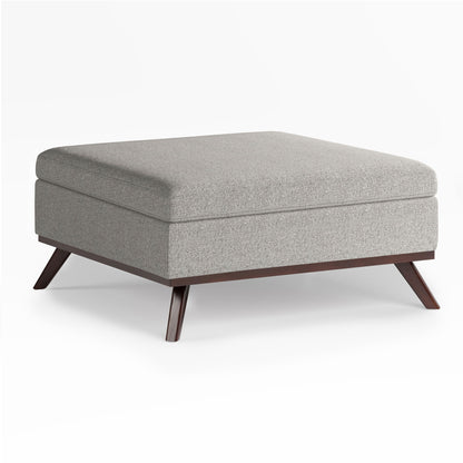 The Owen Multifunctional Square Coffee Table Storage Ottoman features a light gray upholstered, cushioned top and angled dark wood legs, shown against a white background.