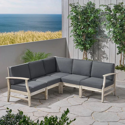 The Perla - 5 Pieces Sofa Set features gray cushions and a light wood frame, set on a stone patio with green plants, offering views of tall grass and the ocean beneath a blue sky.