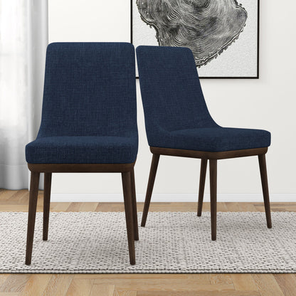 A set of two Kate Mid-Century Modern Dining Chairs with dark blue upholstery and dark wood legs sit on a light textured rug in a modern room featuring wood flooring, a white curtain, and abstract black-and-white wall art.