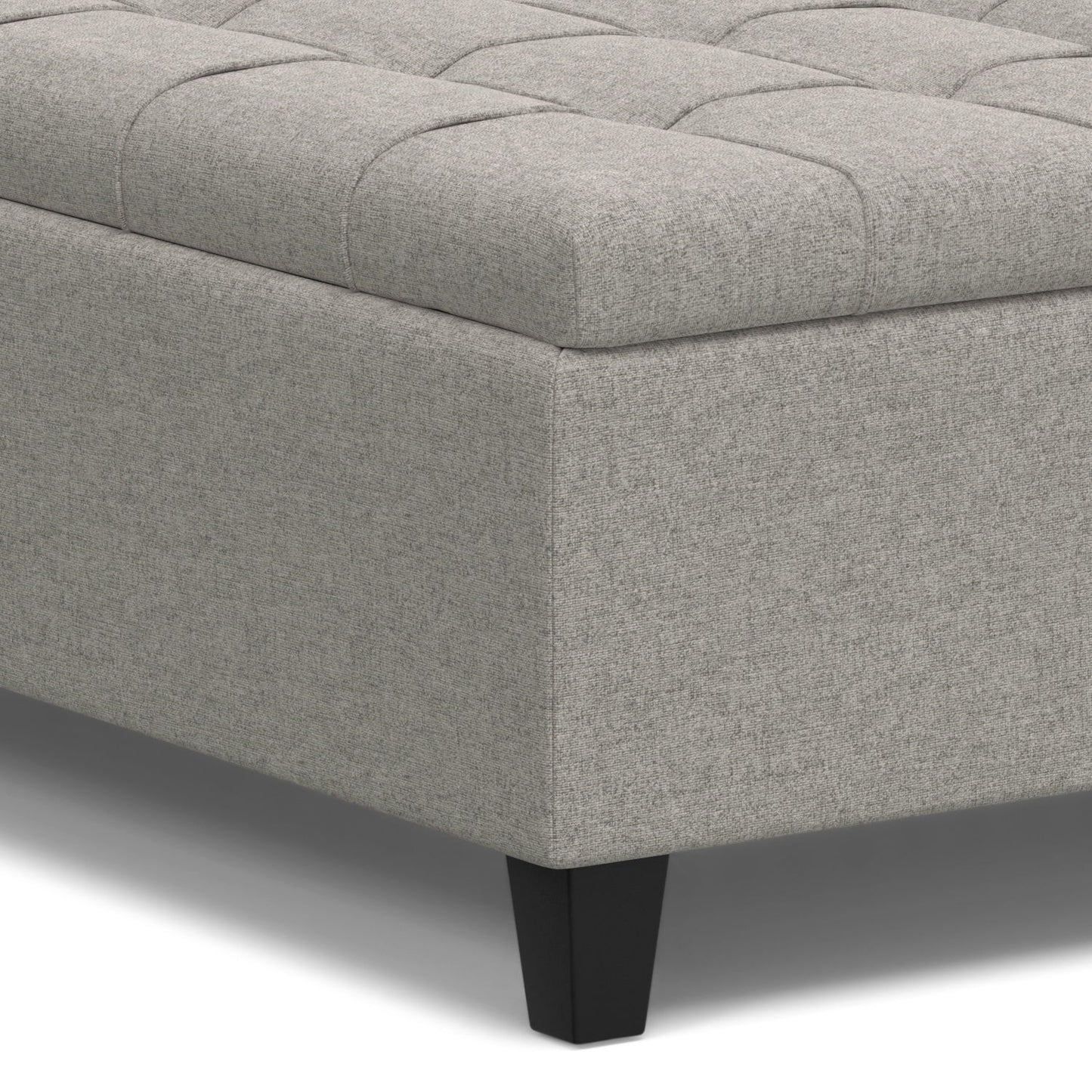 A close-up of the Harrison Upholstered Coffee Table Storage Ottoman shows its light gray tufted top and dark, tapered legs.