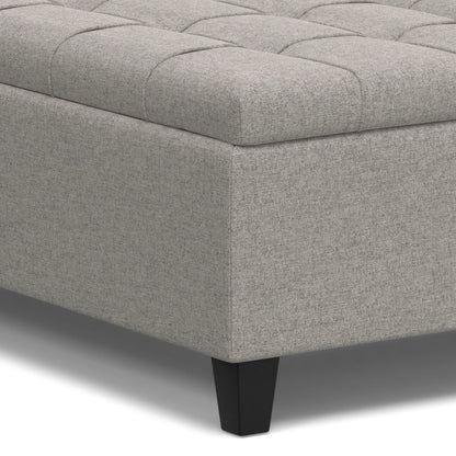 A close-up of the Harrison Upholstered Coffee Table Storage Ottoman shows its light gray tufted top and dark, tapered legs.