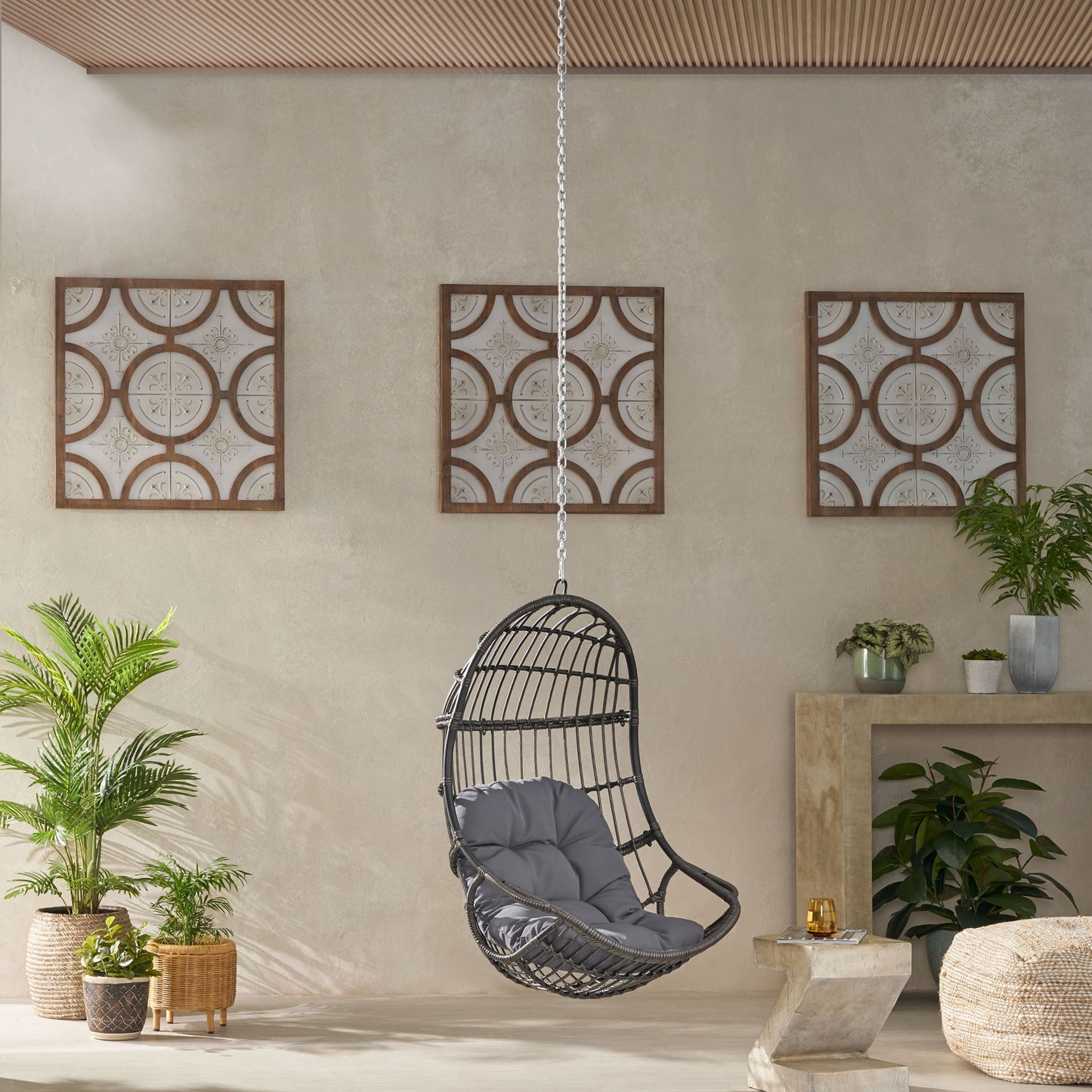 A modern room features the Rustic Hanging Basket Chair With Cushion, black wicker with a gray cushion, surrounded by potted green plants. Three framed panels hang on a beige wall behind, beneath a wooden ceiling.