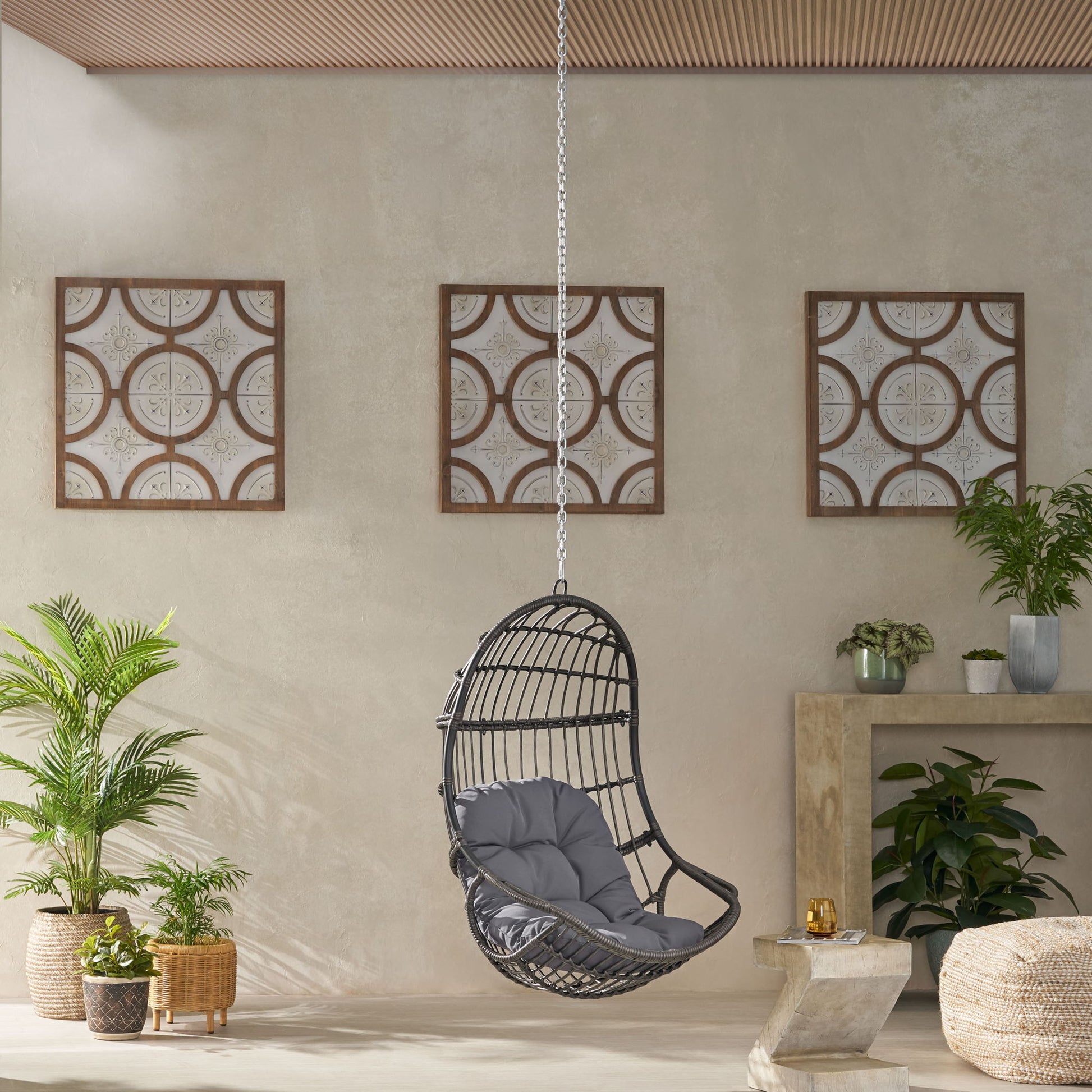 A modern room features the Rustic Hanging Basket Chair With Cushion, black wicker with a gray cushion, surrounded by potted green plants. Three framed panels hang on a beige wall behind, beneath a wooden ceiling.