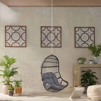 A modern room features the Rustic Hanging Basket Chair With Cushion, black wicker with a gray cushion, surrounded by potted green plants. Three framed panels hang on a beige wall behind, beneath a wooden ceiling.