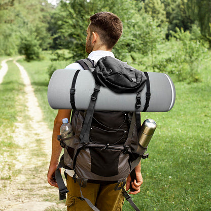 A man with short hair and a beard hikes through green grass and trees, carrying a large black backpack with a Memory Foam Camping Mattress (Portable Comfort With Water Resistant Cover), plus metal and plastic bottles.