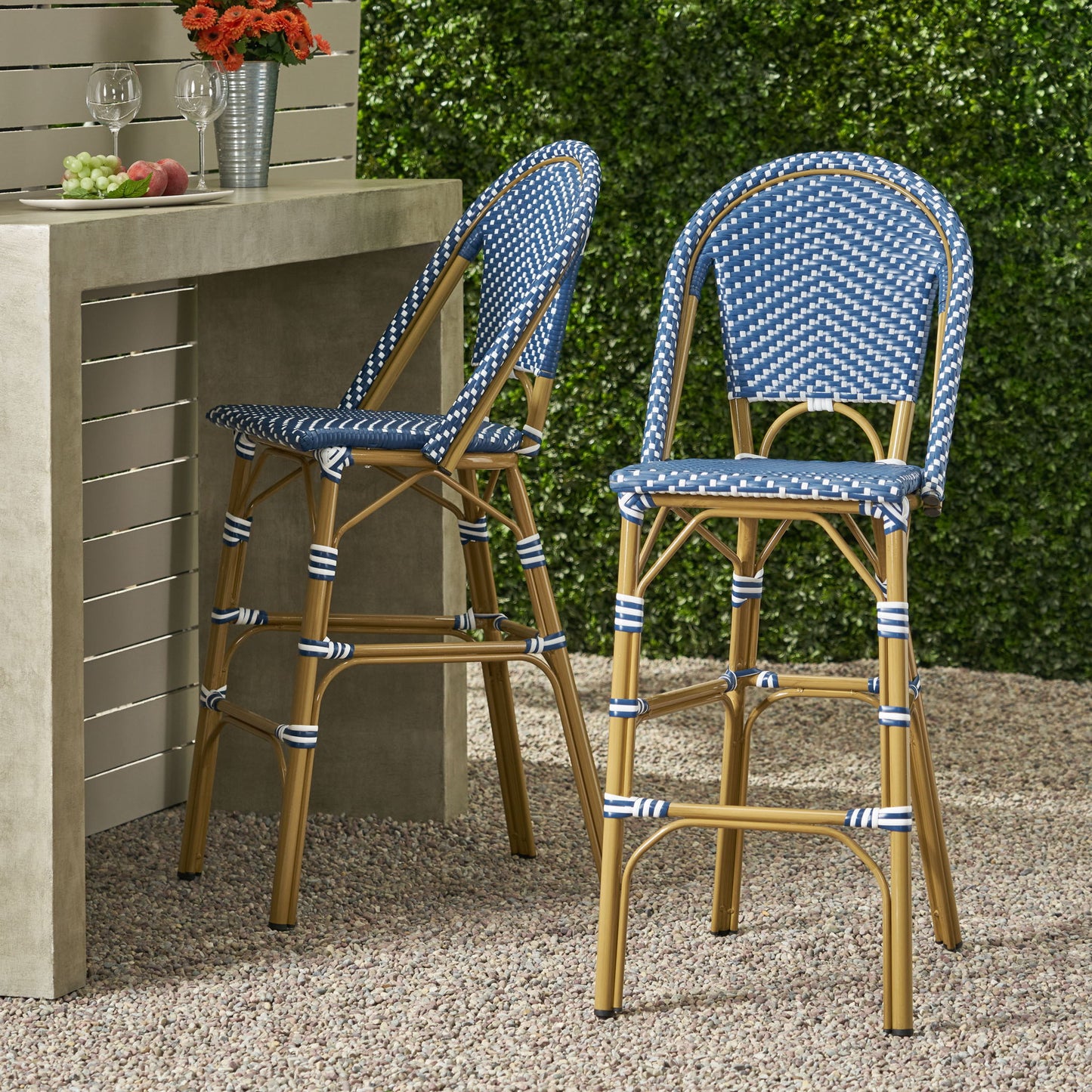 Two Outdoor PE Rattan French Barstools with blue and white weaving and tan legs sit by a concrete outdoor bar table, topped with two glasses of wine, an apple, and grapes. Greenery and gravel complete the scene.
