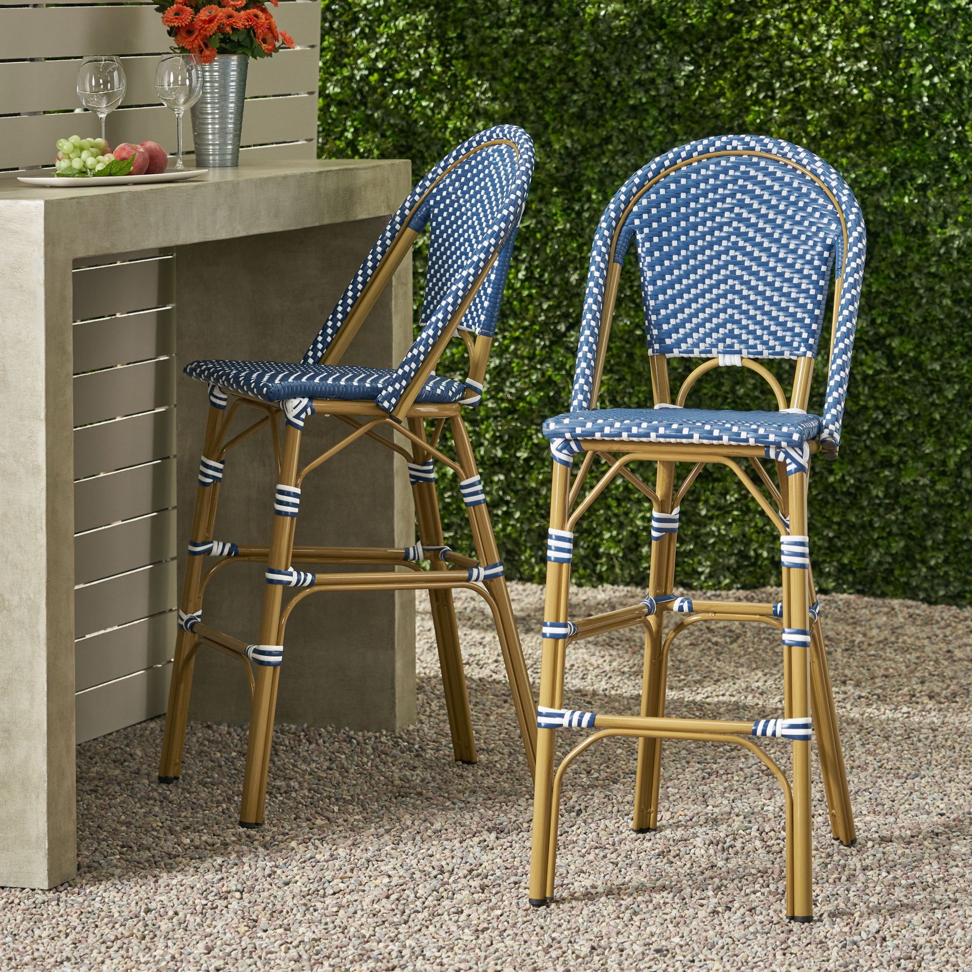 Two Outdoor PE Rattan French Barstools with blue and white weaving and tan legs sit by a concrete outdoor bar table, topped with two glasses of wine, an apple, and grapes. Greenery and gravel complete the scene.