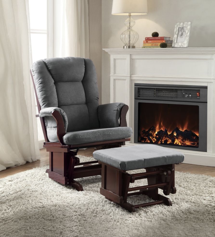 A cozy living room features the Adeliza Microfiber Glider Chair with Ottoman in gray on a plush rug, next to a lit fireplace with a white mantel, lamp, books, and decor. Sunlight filters through curtained windows.