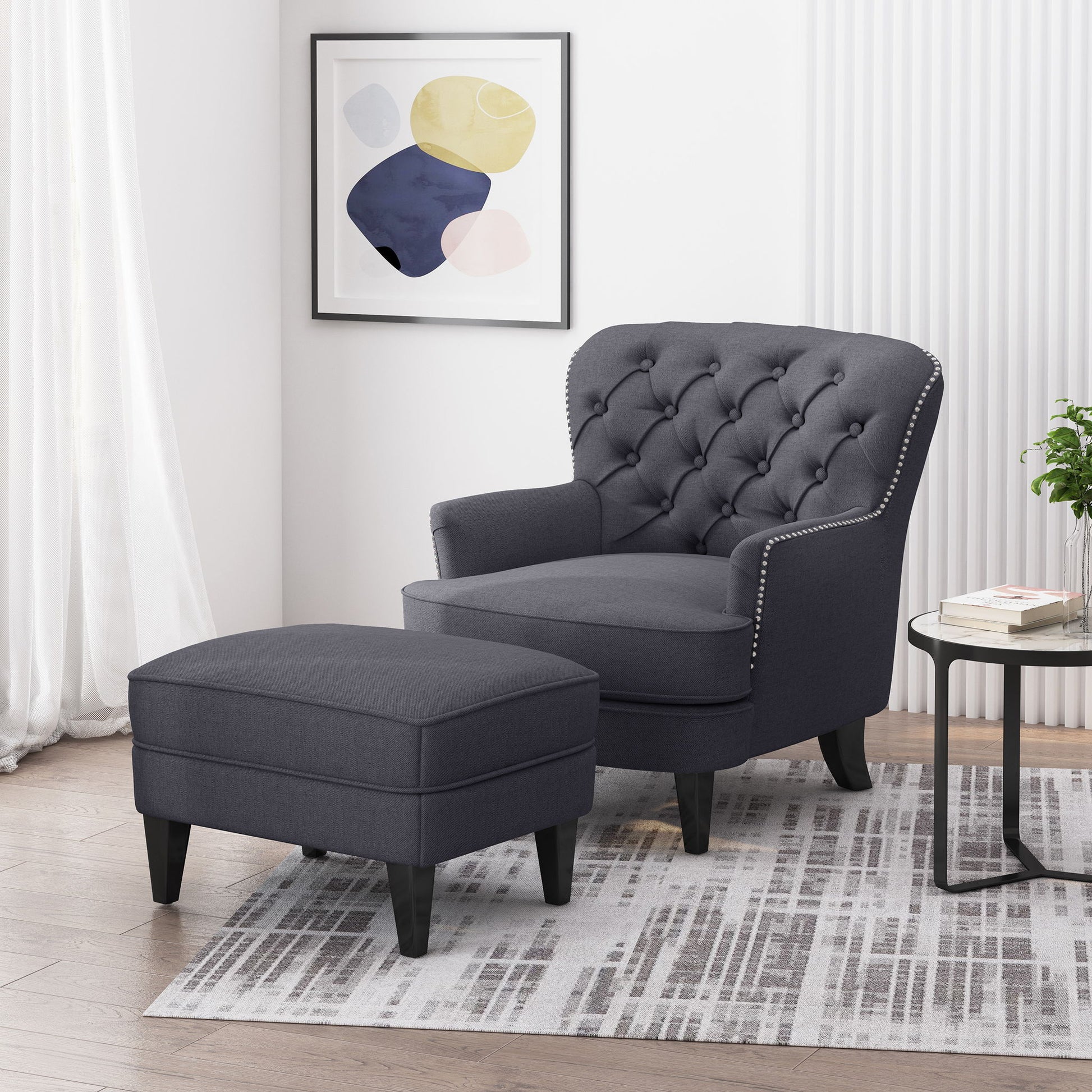 The Enchante Living Room Set with a tufted back club chair and ottoman in dark gray rests on a patterned rug, paired with a round side table holding books and a plant. A modern abstract artwork hangs above.