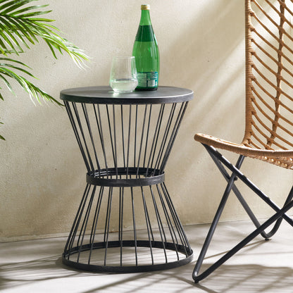 The Lassen - Modern Outdoor Side Table With Hourglass Design holds a green glass bottle and a drinking glass next to a woven chair and leafy plant in a bright, sunlit room.