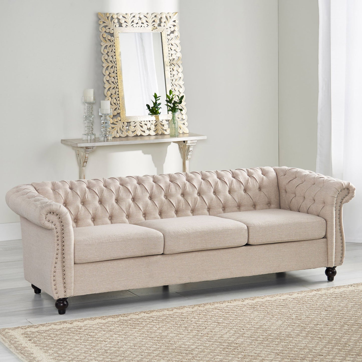 A Chesterfield Style 3 Seater Sofa with deep button tufting and rolled arms is featured in a bright living room, paired with a decorative mirror, shelf with candles and green plants on the white wall, and a light woven rug on the floor.