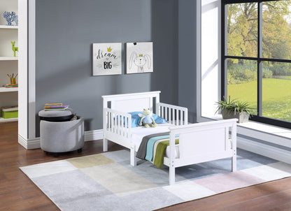 A modern children's bedroom features the Connelly - Reversible Panel Toddler Bed in white, colorful striped bedding, a stuffed animal, wall art, gray ottoman, bookshelf, large window, plant, gray walls, and a pastel area rug.
