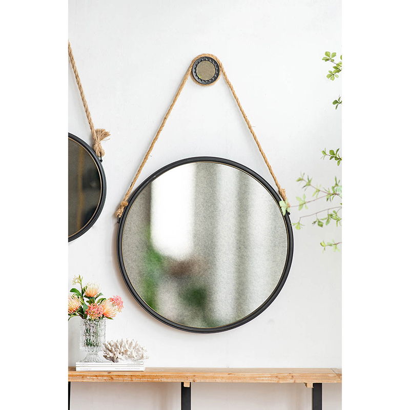 The Handsome Cleveland Mirror with Rope Strap, featuring a round metal frame, hangs on the wall. Below it, a wooden table displays flowers in a glass vase and coral decor. Green branches are visible to the right.