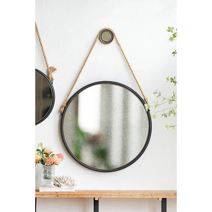 The Handsome Cleveland Mirror with Rope Strap, featuring a round metal frame, hangs on the wall. Below it, a wooden table displays flowers in a glass vase and coral decor. Green branches are visible to the right.