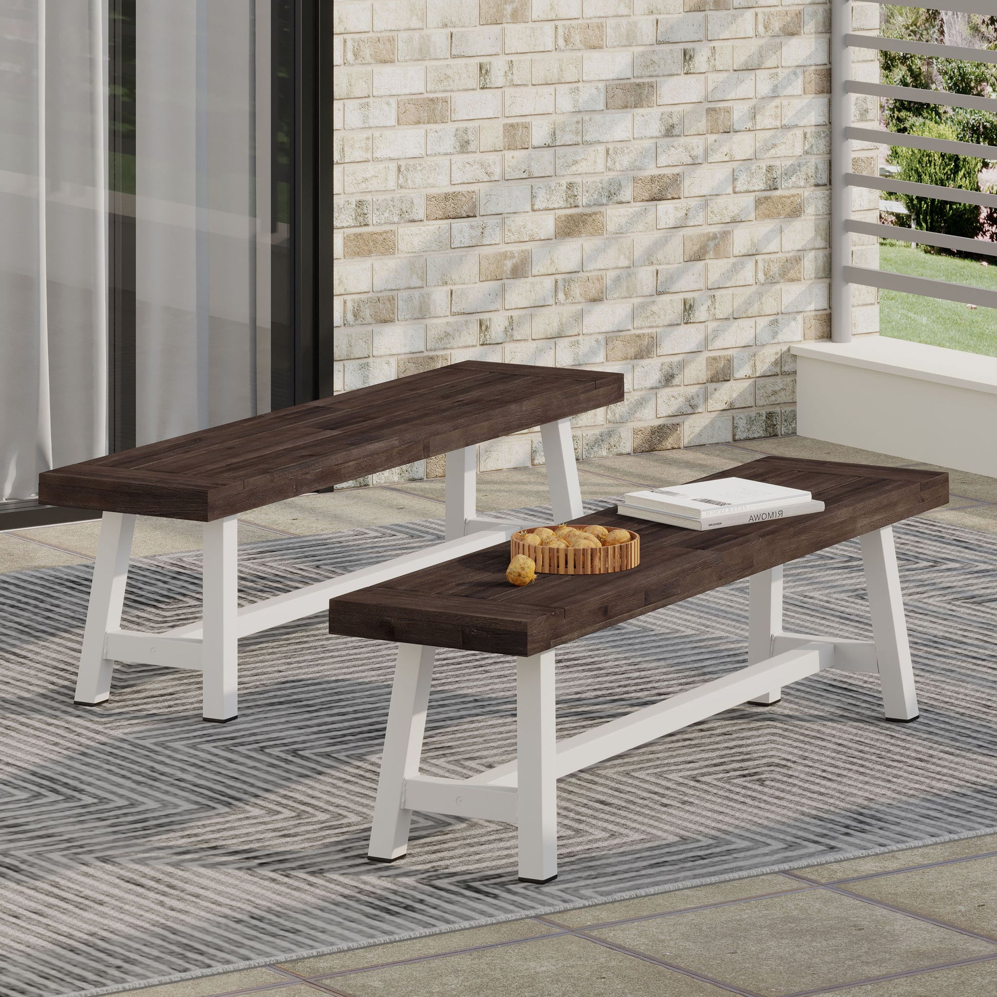 Two Tranquilizer outdoor acacia wood benches with slat panel design, featuring dark brown tops and white legs, sit on a striped rug. One holds a cutting board and book in a modern patio setting with a light brick wall.