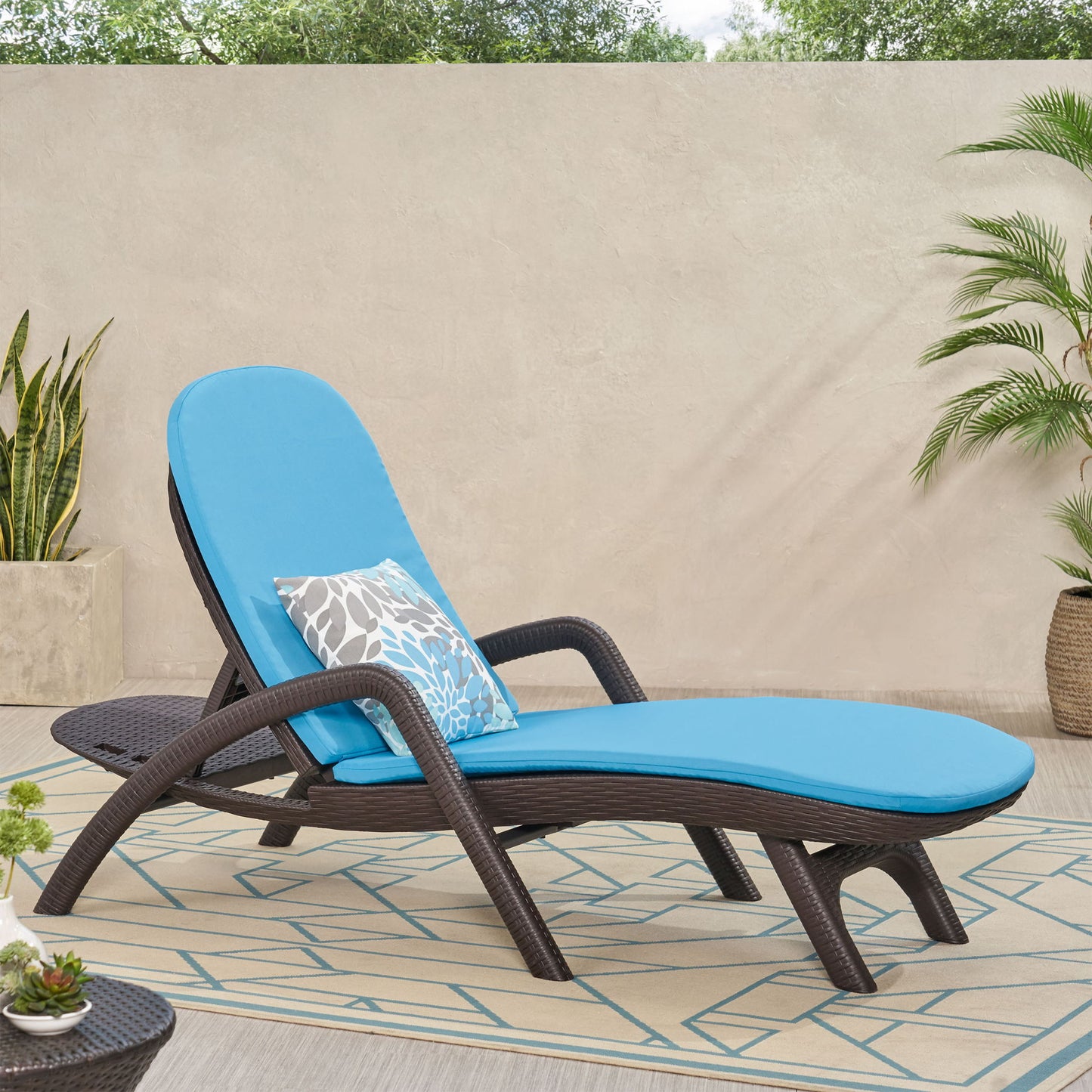 The Mikael Chaise Lounge With Cushion, featuring a bright blue cushion and patterned pillow, is placed on an outdoor rug among potted plants and a concrete wall, with lush greenery in the background.