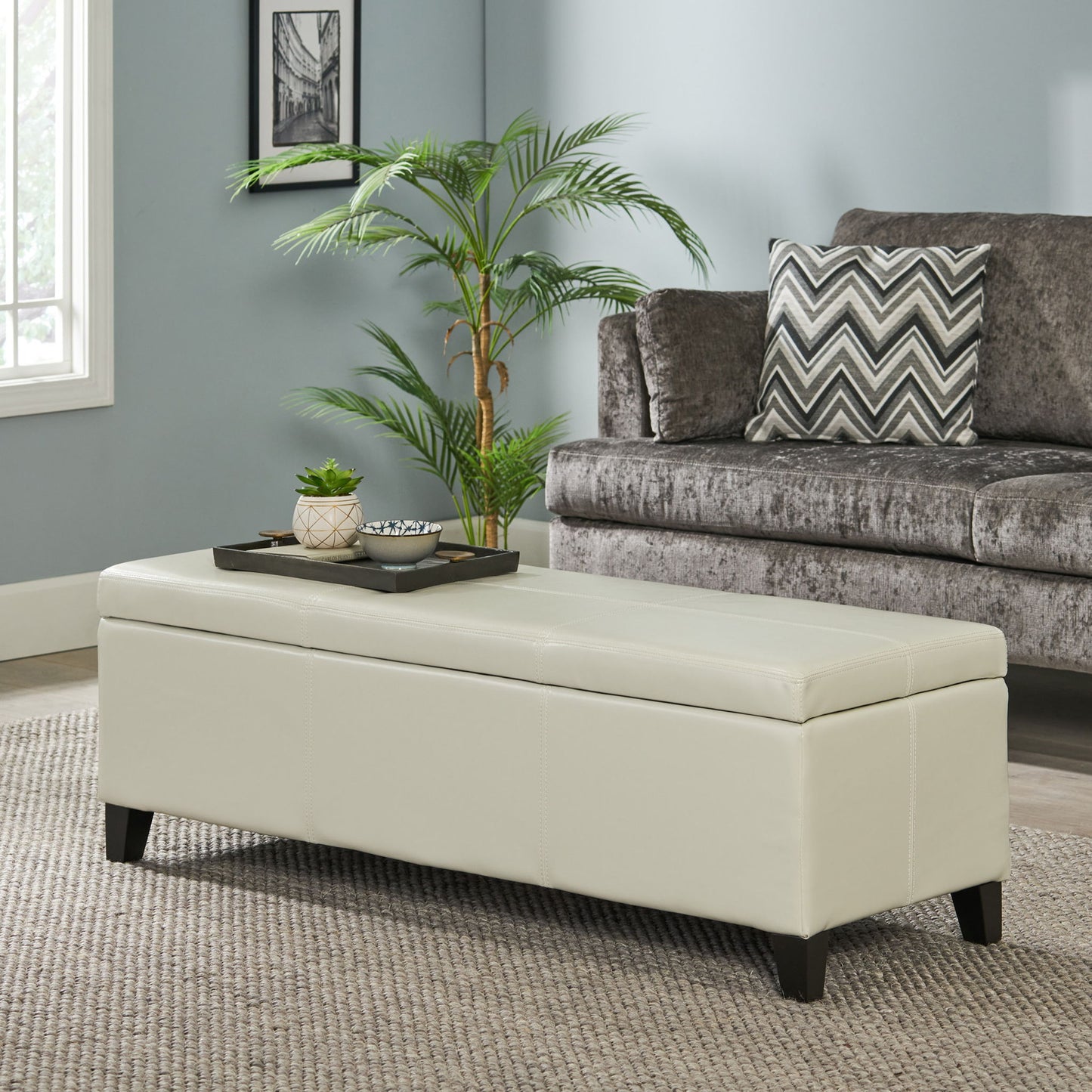 The Glouster - Contemporary Storage Ottoman, upholstered in beige, sits on a textured rug in the living room. A tray with small plants and decor rests on top, while a gray sofa with chevron pillows and a potted plant are in the background.