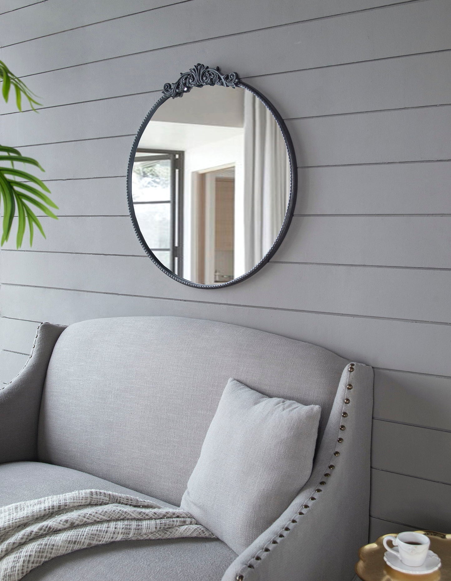 A Classic Design Mirror With Baroque Inspired Frame hangs on a gray shiplap wall above a light gray upholstered sofa with cushion and blanket. Nearby are a potted plant and a small side table with a cup.