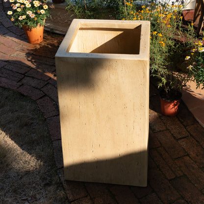 A spacious, square beige Modern Outdoor Planter sits on a brick patio, surrounded by potted yellow and white flowers in sunlight and partial shade.