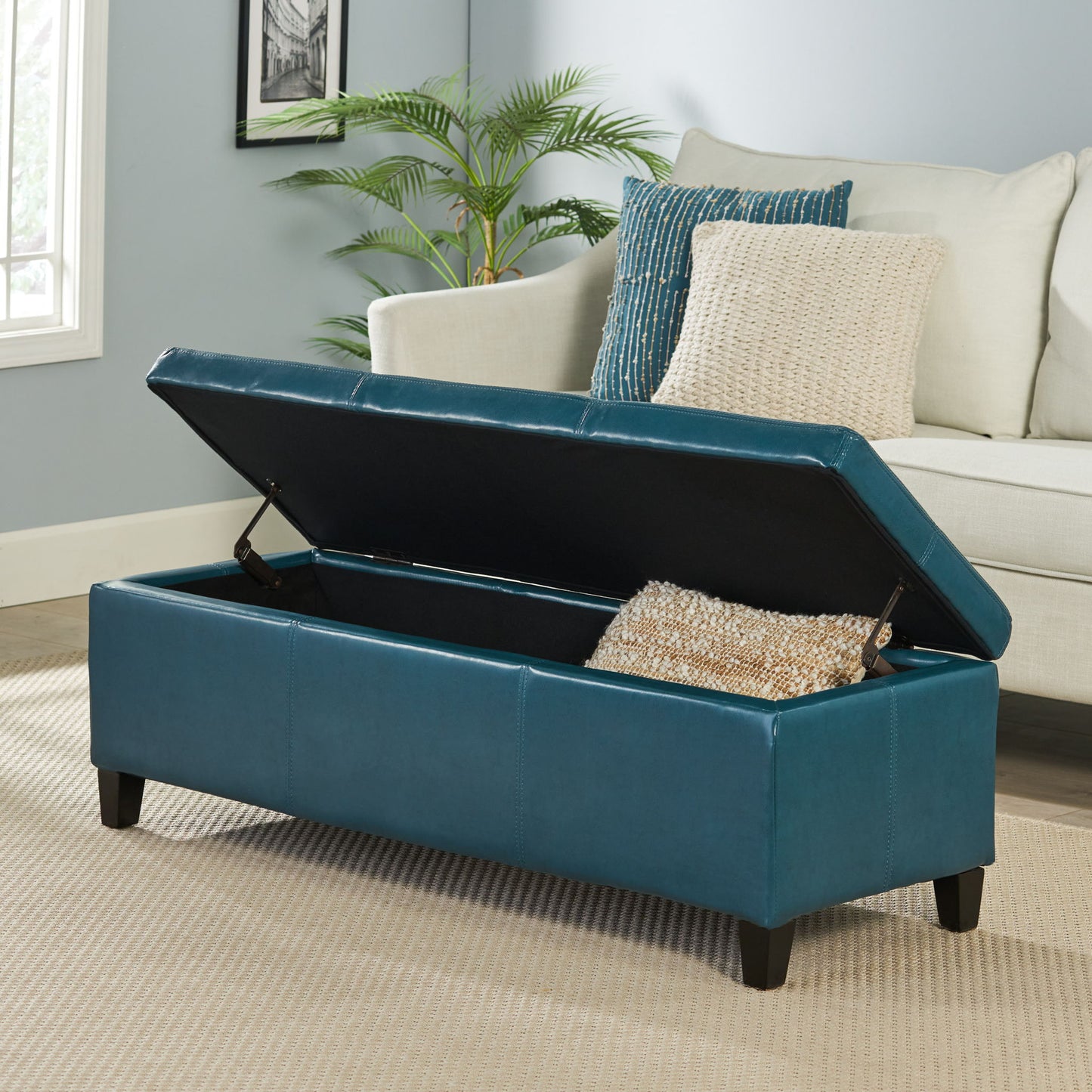 The Glouster - Contemporary Storage Ottoman with its lid open sits on a beige rug in a living room, showing a decorative pillow inside. In the background are a light-colored sofa with textured cushions and a potted plant.