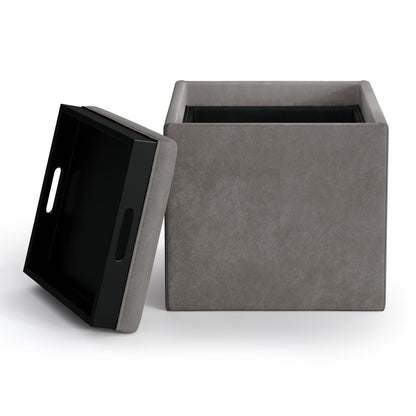 The Rockwood Upholstered Cube Storage Ottoman With Tray is a square, gray ottoman with a removable lid featuring cutout handles and a tray. When lifted, the lid reveals a black storage compartment inside. Shown against a plain white background.