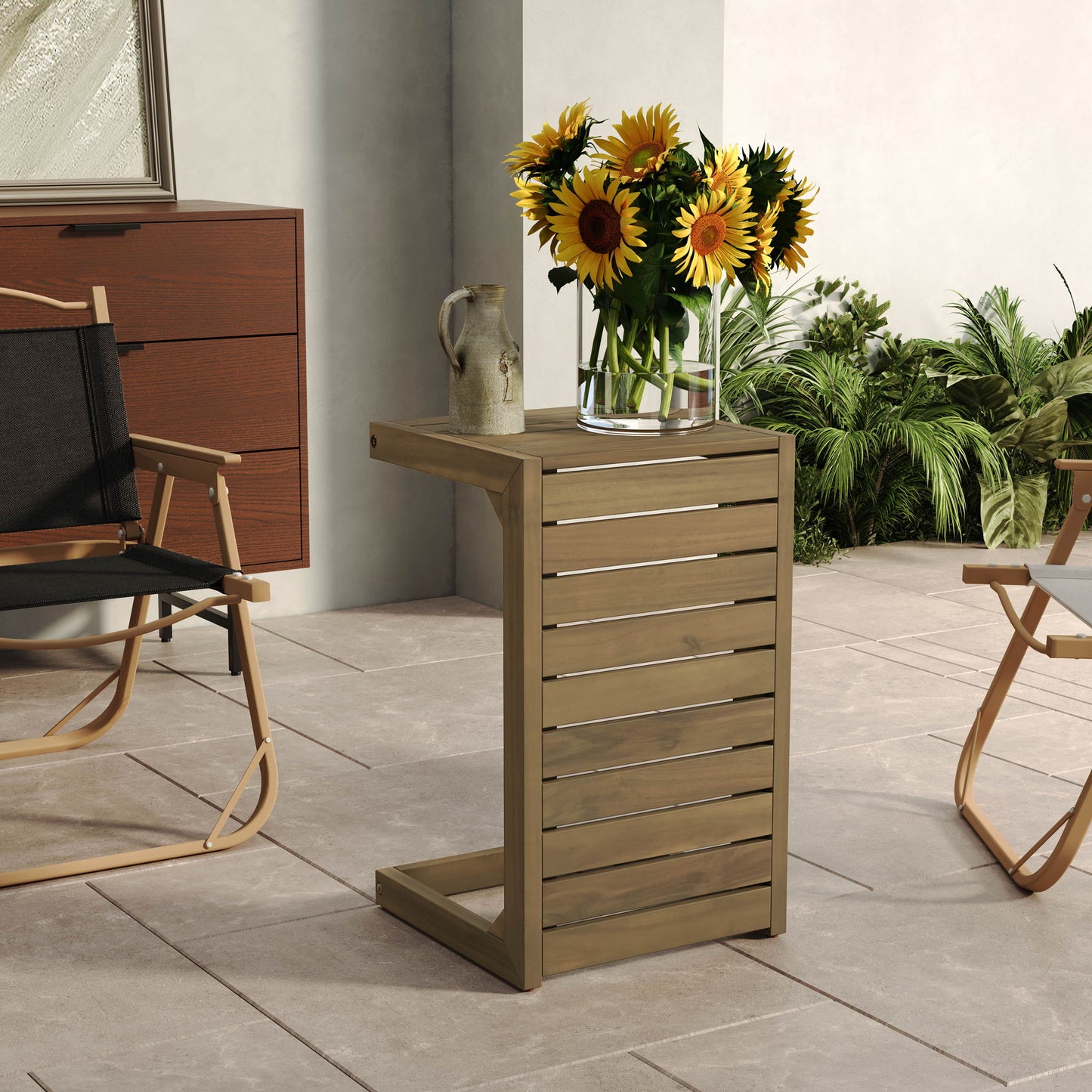 An Aluminum Outdoor C-Shaped Side Table for Patio displays a clear vase of sunflowers and a ceramic jug on a tiled patio, amidst chairs, greenery, and a brown cabinet.