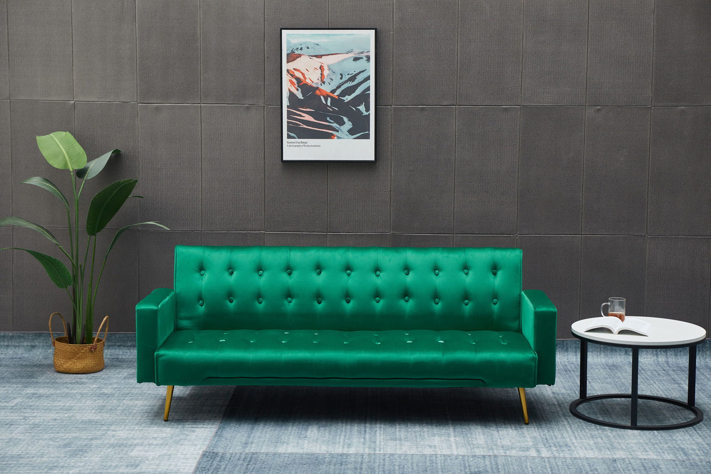 A modern living room features the Comfort Compact Design Sofa Bed in green, a potted plant on the left, a round white coffee table with an open book and glass, and framed abstract art on a gray textured wall.