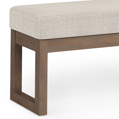 A close-up of the Milltown Upholstered Ottoman Bench, showcasing its light beige cushion and rectangular wooden frame with a brown finish. The design is simple and contemporary.