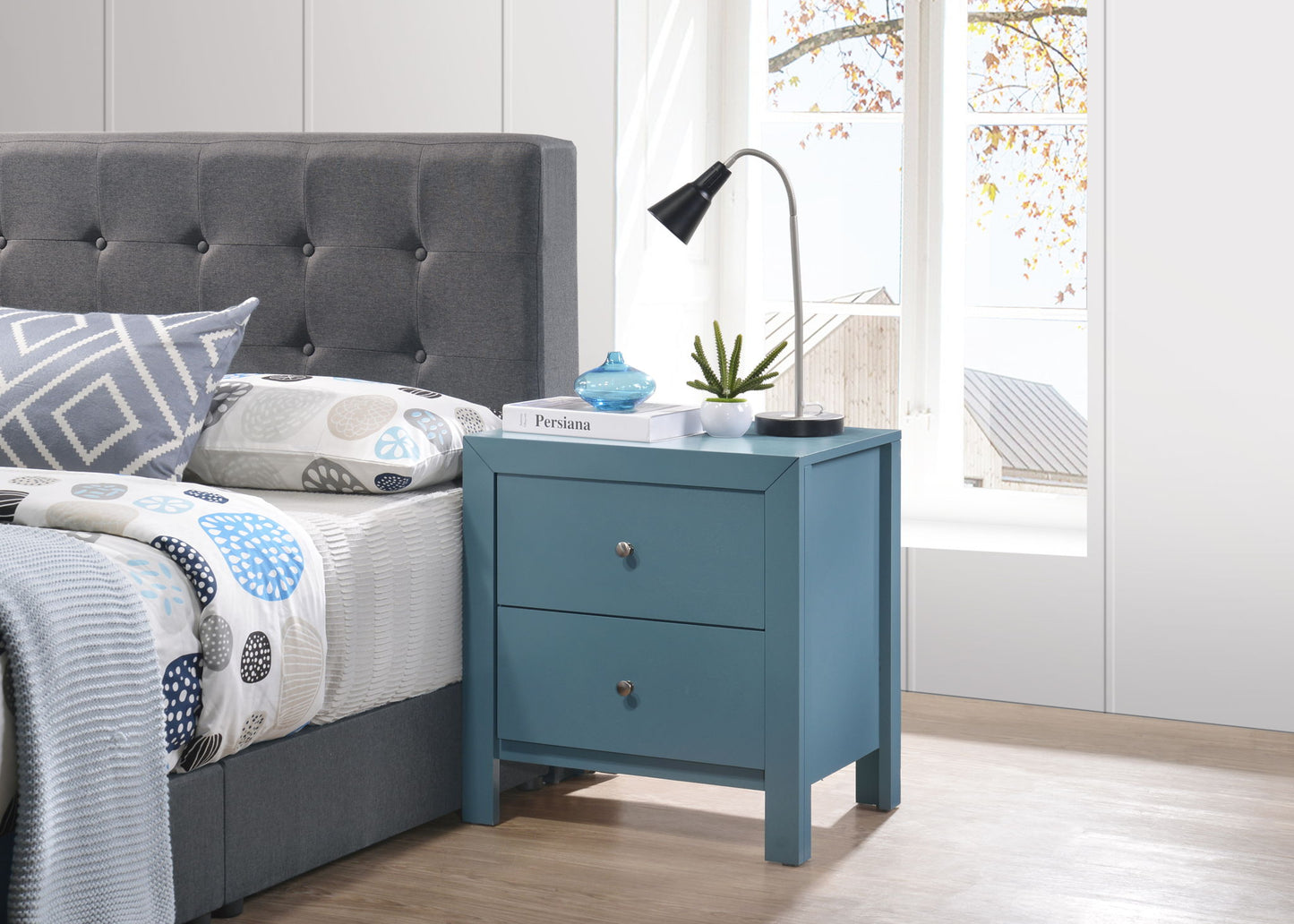 A modern bedroom with a gray tufted bed, patterned bedding, and a Charming Nightstand With Drawers holding a lamp, potted plant, book, and blue jar, set by a large window showing autumn leaves.