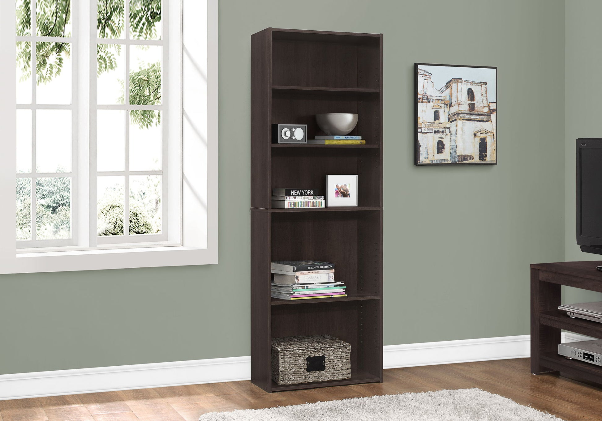 A 6-tier transitional bookcase stands by a window against a light green wall, holding books, decor, and a radio. Nearby are artworks and a TV.