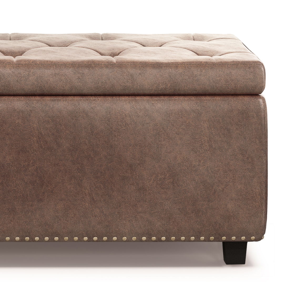 The Hamilton Upholstered Storage Ottoman features brown faux leather, tufted details, decorative nailhead trim at the base, and short black wooden legs. Only part of the ottoman appears against a white background.