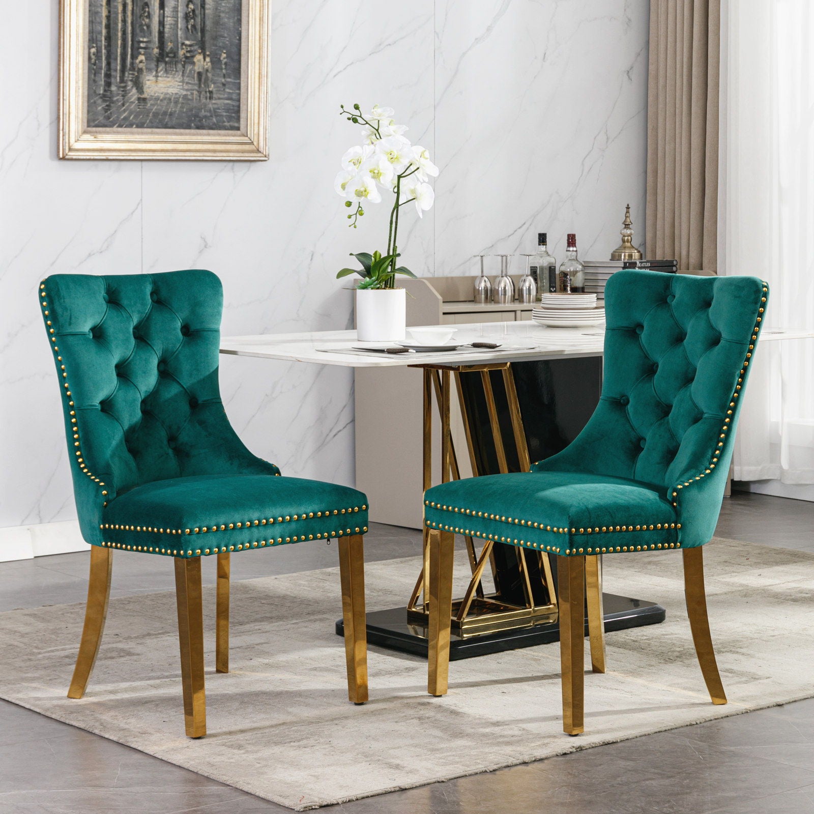 Two Nikki modern tufted green velvet dining chairs with golden stainless steel legs and nailhead trim stand before a marble-top table set with glassware and a white orchid. The stylish room features modern decor, a painting, and a large window.