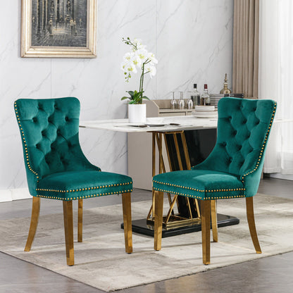 Two Nikki modern tufted green velvet dining chairs with golden stainless steel legs and nailhead trim stand before a marble-top table set with glassware and a white orchid. The stylish room features modern decor, a painting, and a large window.