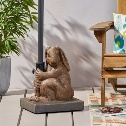 The Hand Crafted Rabbit Umbrella Base With Playful Design in light brown sits beside a wooden chair, potted plant, and colorful rug on a wooden deck, adding decorative charm to your patio setup.