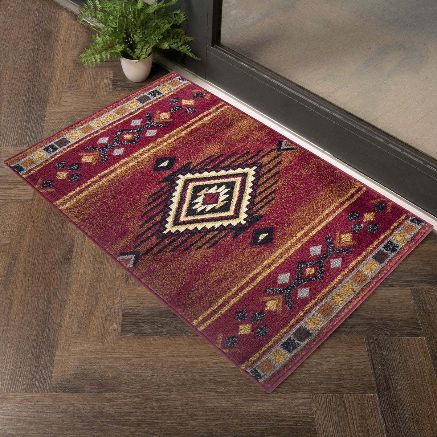 The Tribes - GC_YLS4002 Southwest Area Rug with red and gold geometric patterns lies on a wooden floor near a glass door, complemented by a nearby potted green plant.