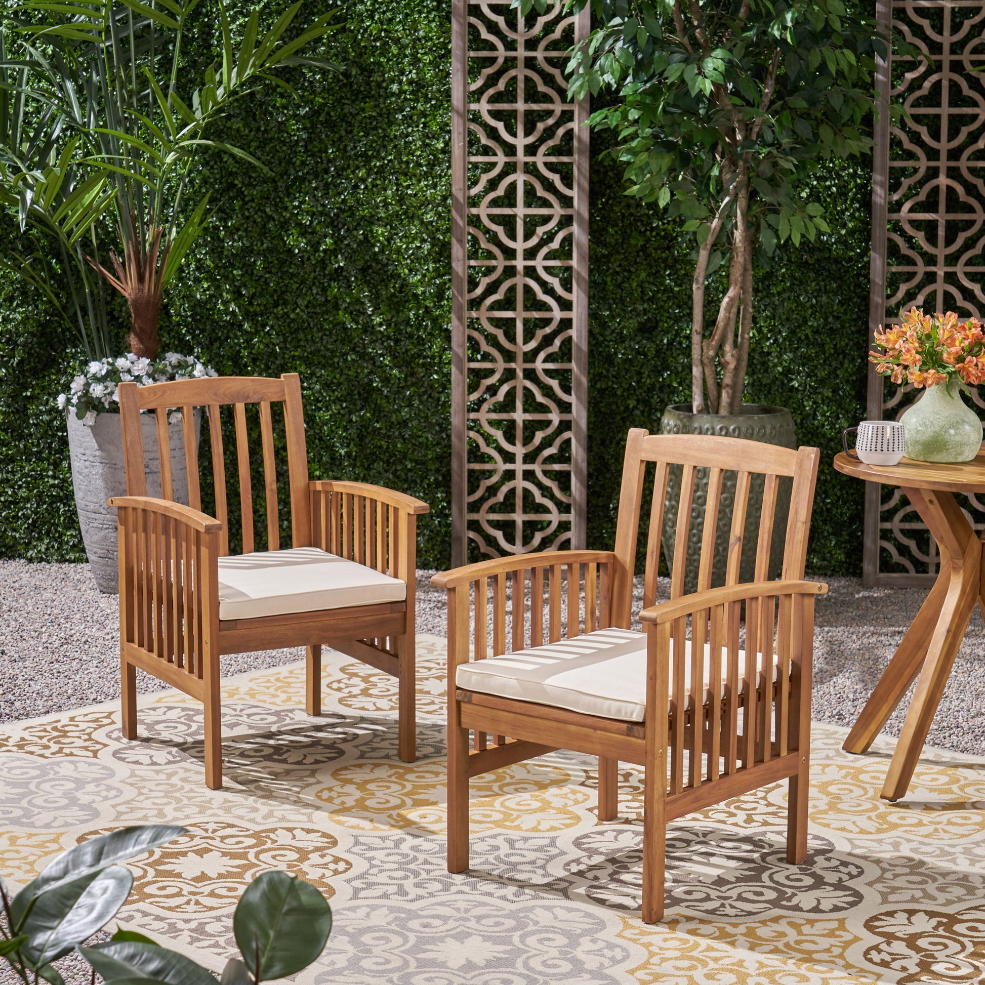 Two Casa Acacia Dining Chairs with cream cushions sit on a patterned outdoor rug in a garden, surrounded by greenery. Nearby, a small round wooden table holds decorative items amidst lush plants and trees.