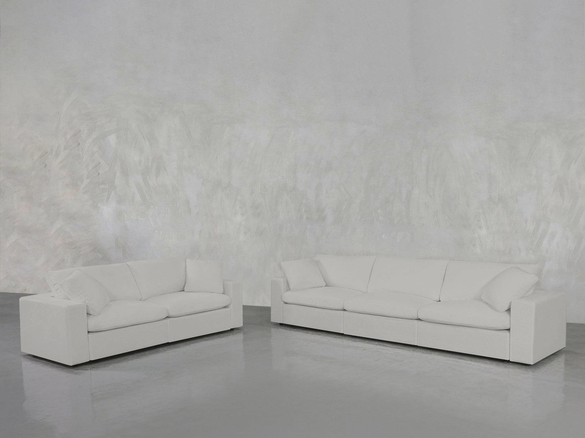 The 3-2 Sofa Set, featuring modern white sofas with clean lines, is arranged in a minimalistic room with a textured light gray wall and a shiny gray floor.