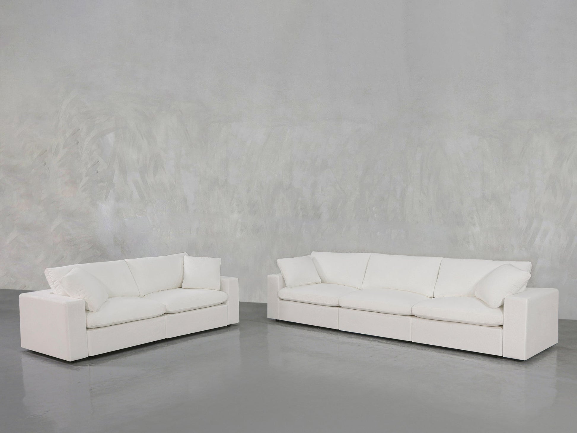 The 3-2 Sofa Set, featuring modern white sofas with plush cushions, is arranged on a shiny gray floor against a textured light gray wall in a minimalistic room.