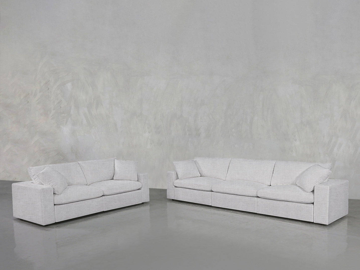 The 3-2 Sofa Set, featuring modern light gray fabric upholstery and large cushions, sits on a smooth gray floor against a textured light gray wall, creating a minimalist and spacious atmosphere.