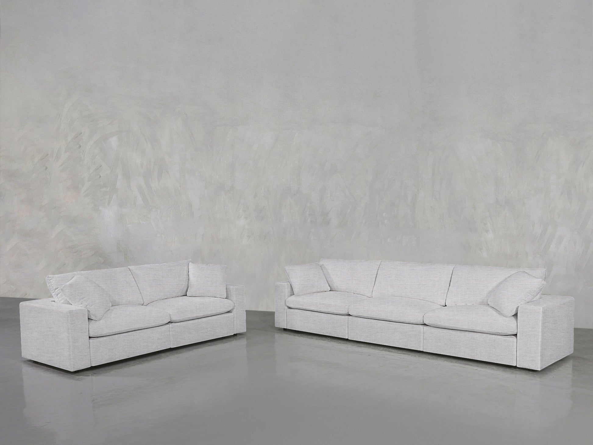 The 3-2 Sofa Set, featuring modern light gray fabric upholstery and large cushions, sits on a smooth gray floor against a textured light gray wall, creating a minimalist and spacious atmosphere.
