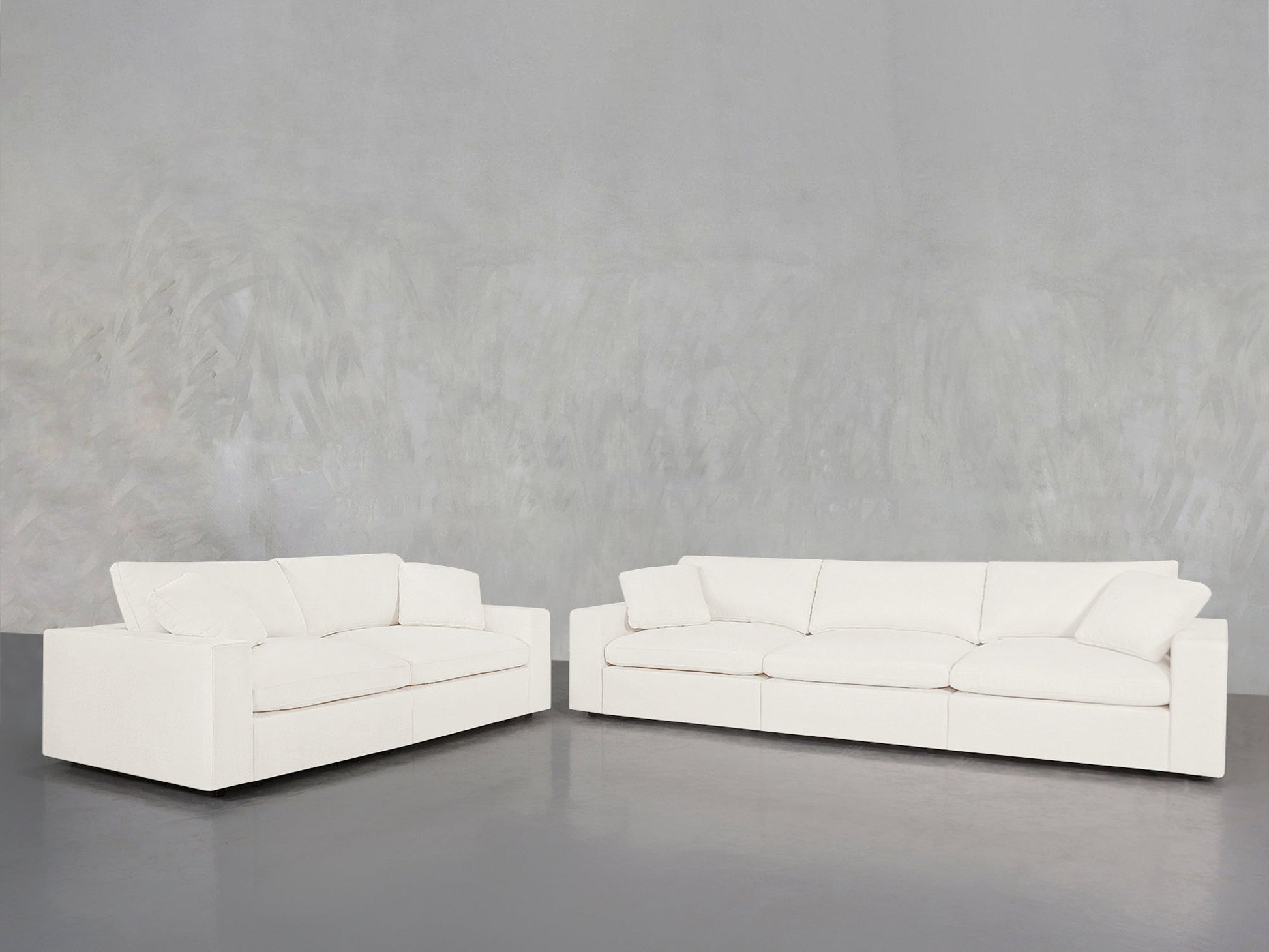 The 3-2 Sofa Set, featuring wide armrests and plush cushions in a modern white design, is arranged in an L-shape against a light gray textured wall on a smooth gray floor.