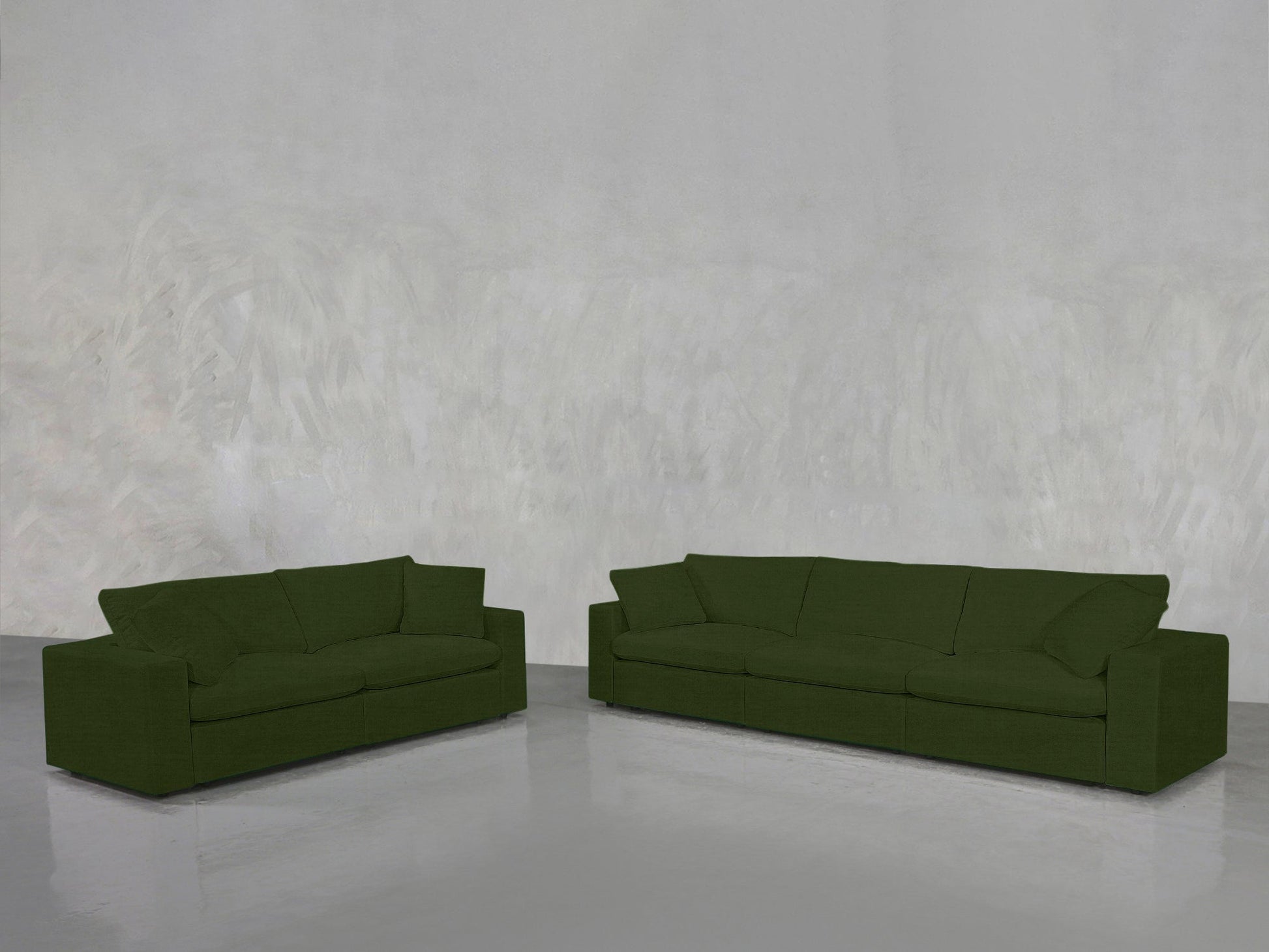The 3-2 Sofa Set, featuring both a smaller and larger green sofa, is arranged on a shiny grey floor against a textured light grey wall in a minimalist, empty room.