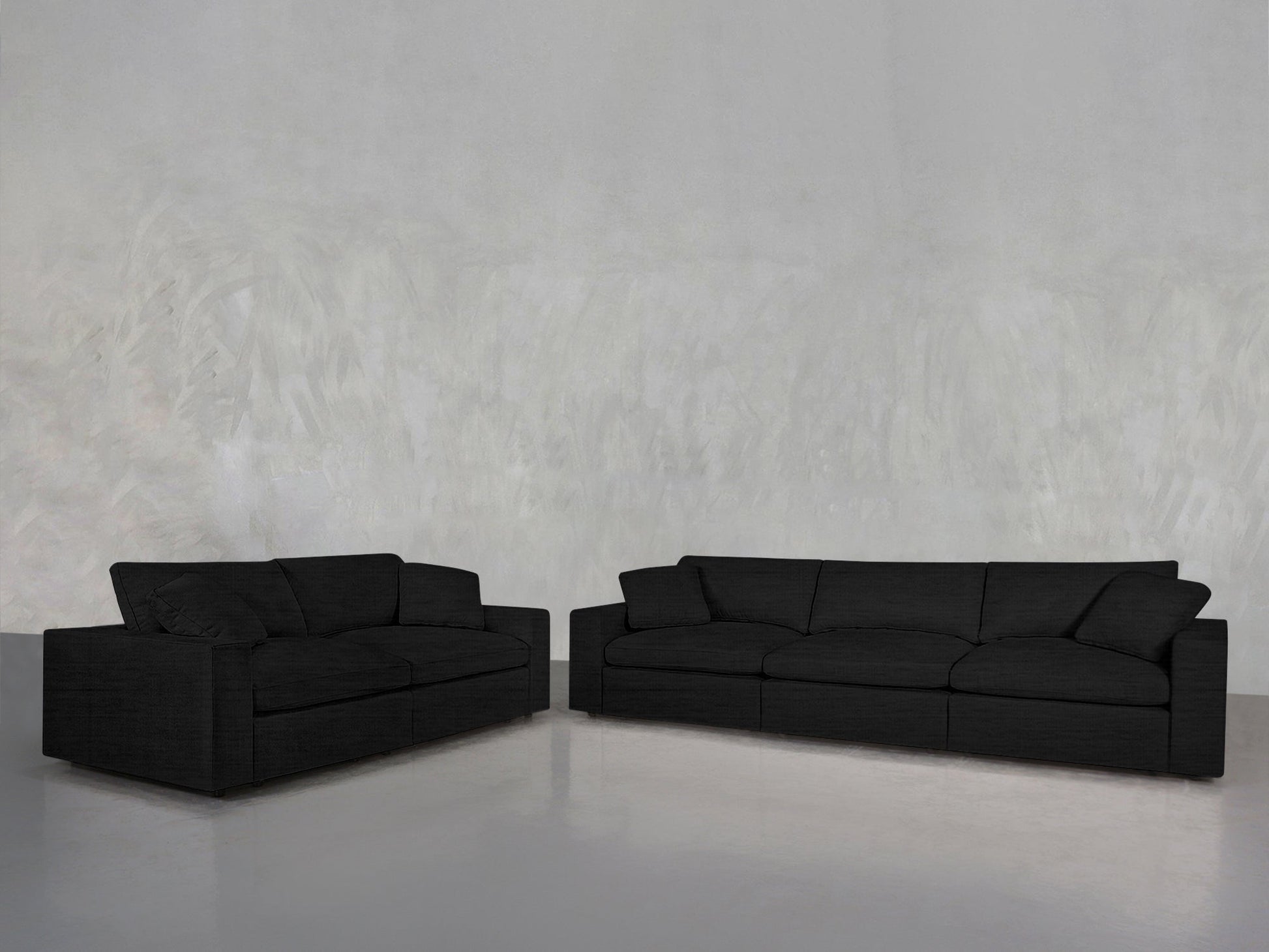 The 3-2 Sofa Set, featuring modern black fabric two- and three-seater sofas, is arranged in an empty room with a light gray textured wall and a smooth gray floor.